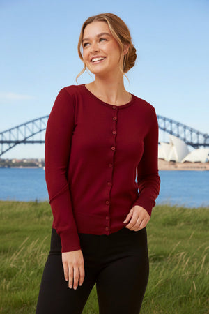 Woman wearing a red cardigan standing in a grassy area with a bridge and sailboat in the background.