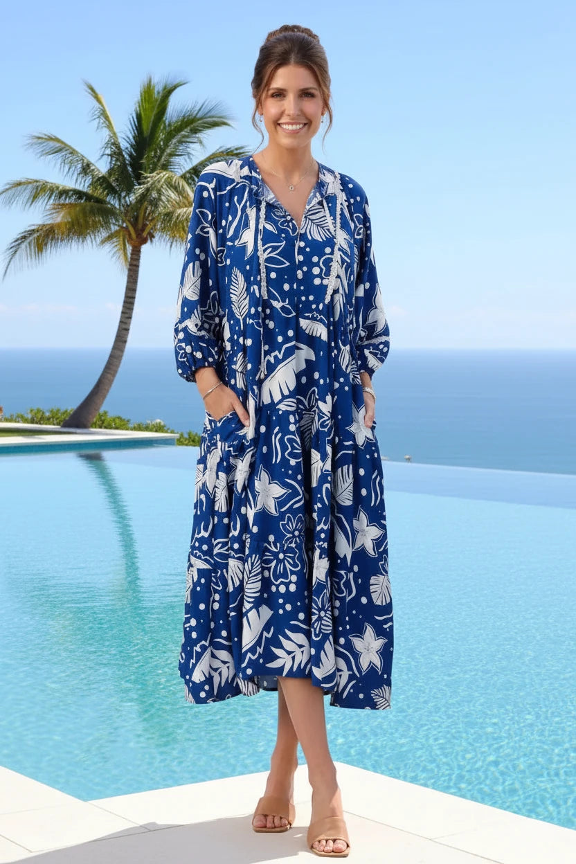 Woman in a blue floral dress standing by a pool with palm trees and ocean in the background