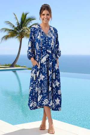 Woman in a blue floral dress standing by a pool with palm trees and ocean in the background