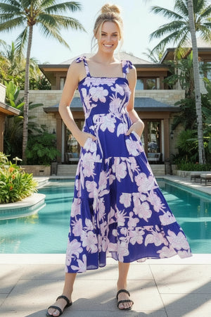 Woman in a blue floral dress standing by a pool with palm trees in the background