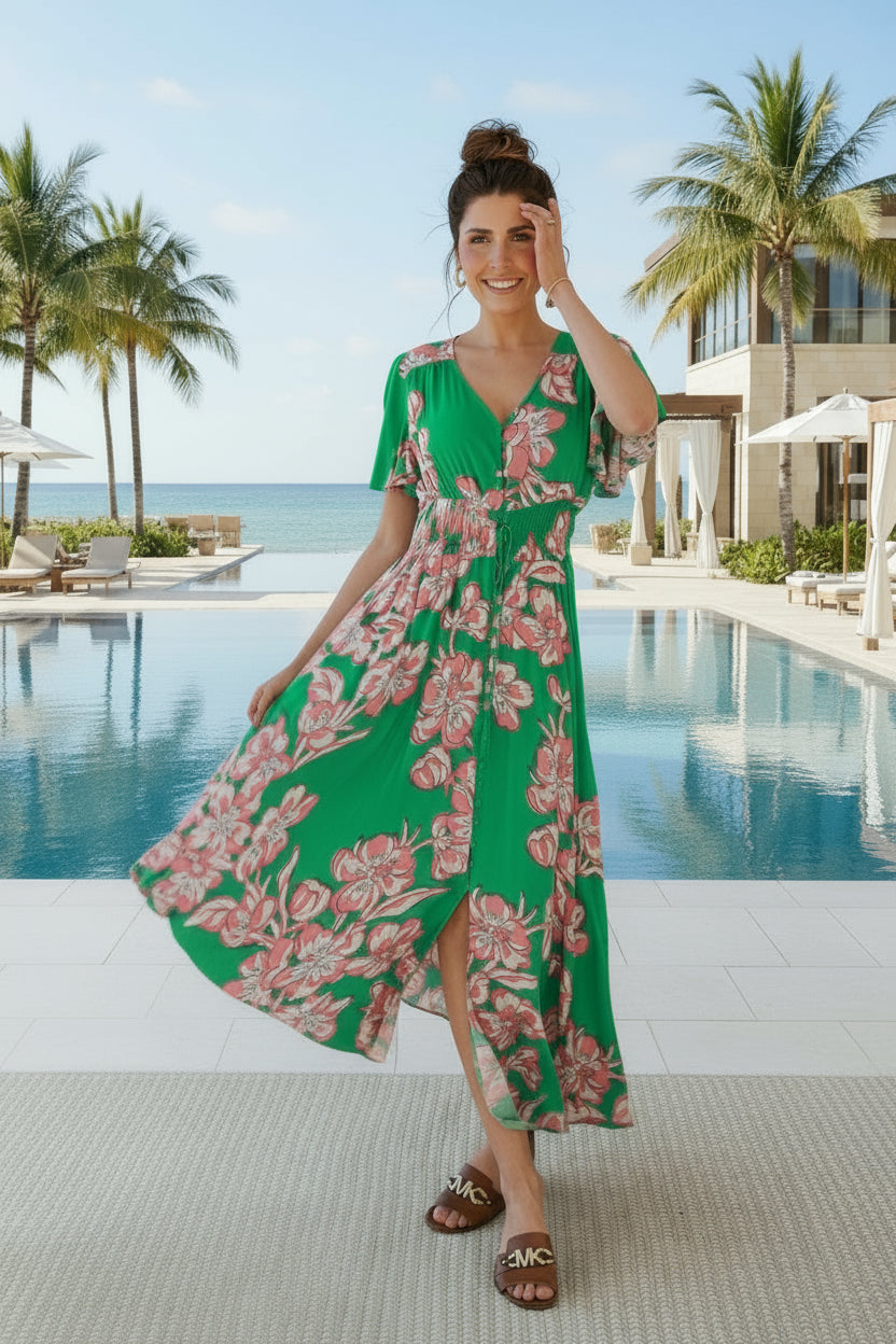 Woman in a green floral dress standing by a pool with palm trees and a building in the background.