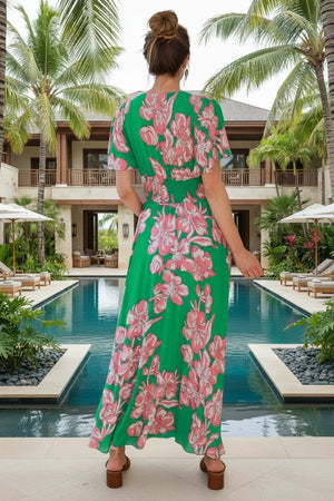 Woman in a green floral dress standing by a pool with palm trees and a building in the background