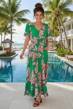 Woman in a green floral dress standing by a pool with palm trees and a building in the background.