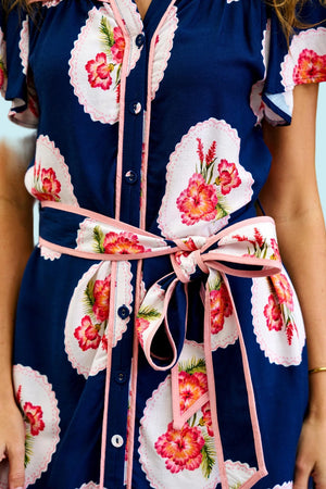 A close-up of a navy blue shirt dress with a pink and white hibiscus floral print, featuring a matching pink tie belt at the waist.