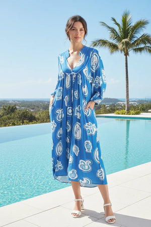 Woman wearing a blue dress with white patterns in an outdoor setting with plants and stones.