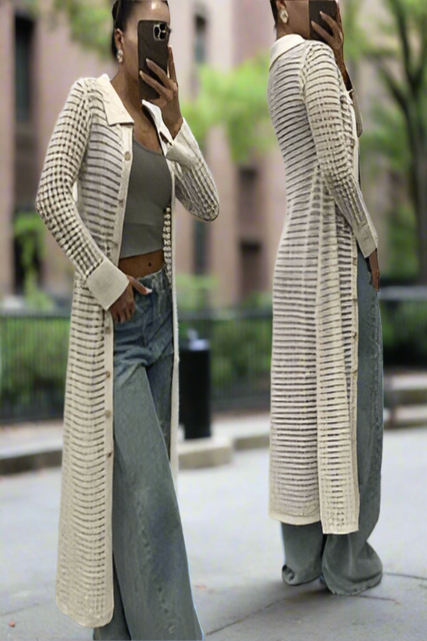 A model wearing a beige long crochet knit maxi dress or jacket with button down collar, over a slip, paired with jeans on a sunny day.