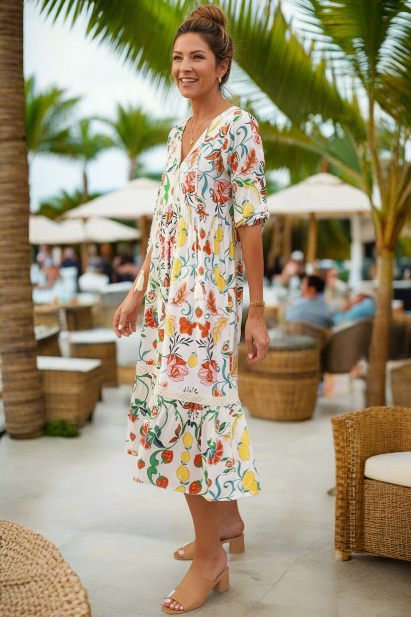Woman in a floral dress standing in a tropical outdoor setting with palm trees and people in the background.