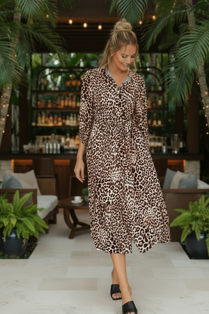 Woman in a leopard print dress standing in an outdoor setting with palm trees and a bar in the background.