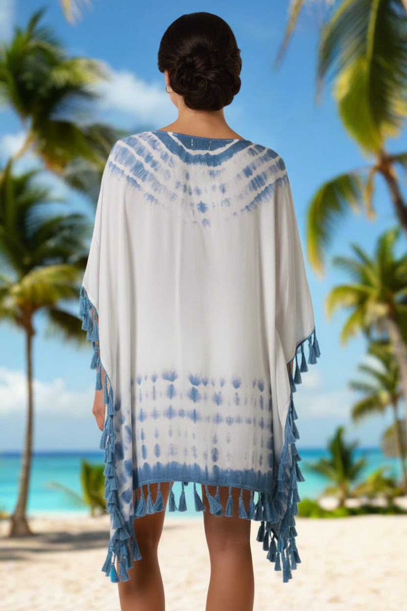 Woman wearing a white and blue patterned cover-up with tassels on a beach with palm trees and blue sky.