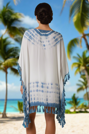 Woman wearing a white and blue patterned cover-up with tassels on a beach with palm trees and blue sky.