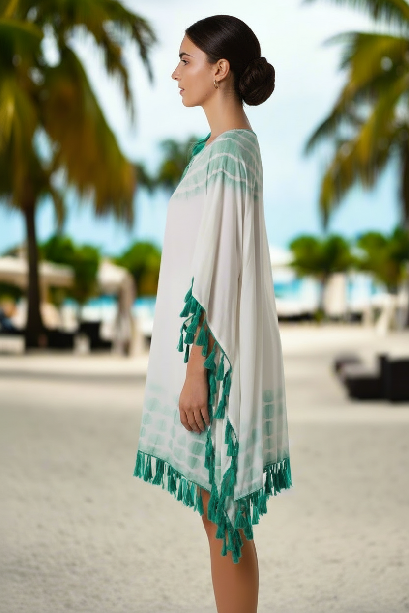 Woman wearing a white and green dress with tassels on a beach with palm trees in the background