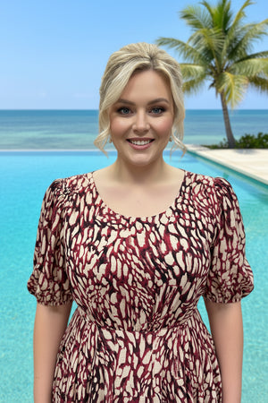 Woman in a patterned dress standing by a pool with palm trees and ocean in the background