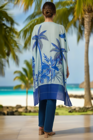 Woman wearing a blue and white kimono-style garment with palm tree design, standing on a wooden deck with palm trees and ocean in the background.