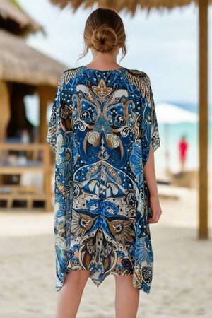 Woman in a blue patterned dress with a straw hat on a beach.