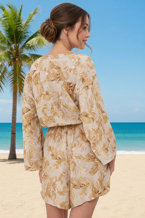 Woman in a beige leaf print playsuit standing on a beach with palm trees and blue sky.