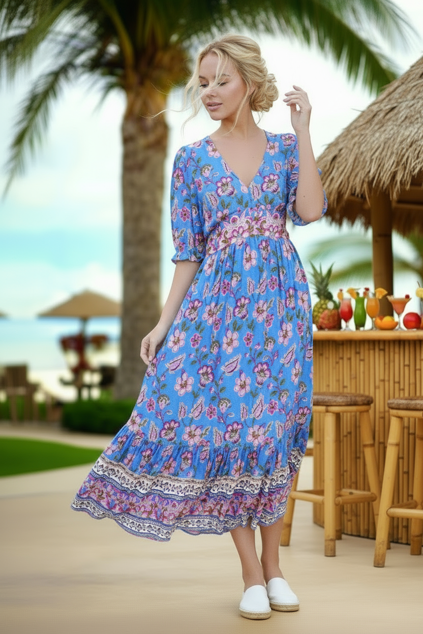 Woman in a blue floral dress standing in a tropical setting with palm trees and a bar.