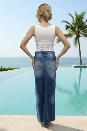 Woman wearing a blue denim maxi skirt standing by a pool with palm trees in the background.