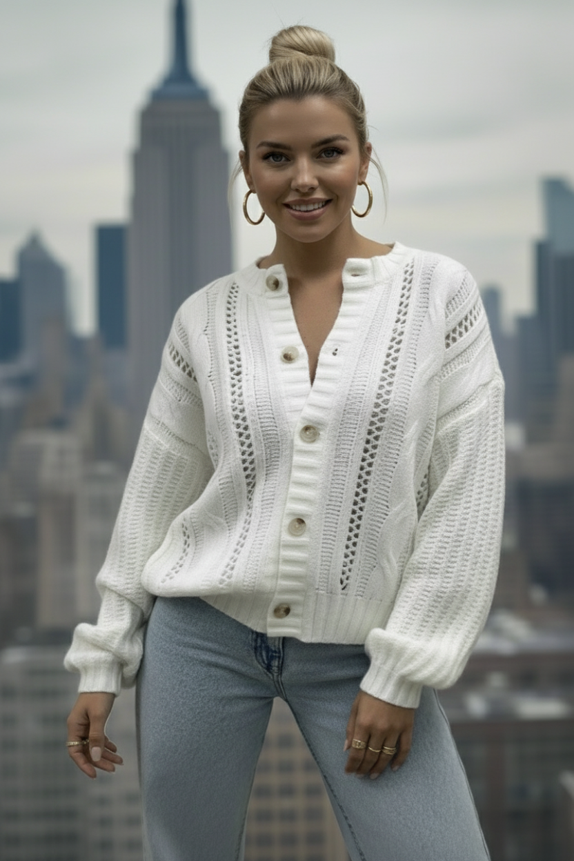 A person wearing a white knit bomber style cardigan with button down front, taking a picture of a cityscape with a smartphone.