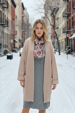 Woman standing on a snowy street wearing a long beige coat, gray dress, and floral scarf.