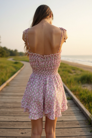 Woman in a pink floral dress standing on a wooden boardwalk by a beach at sunset.