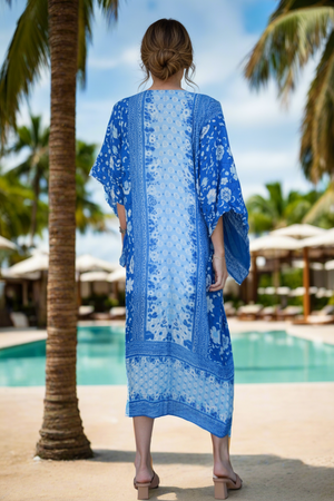 Woman in a blue patterned dress standing by a pool with palm trees and umbrellas in the background.