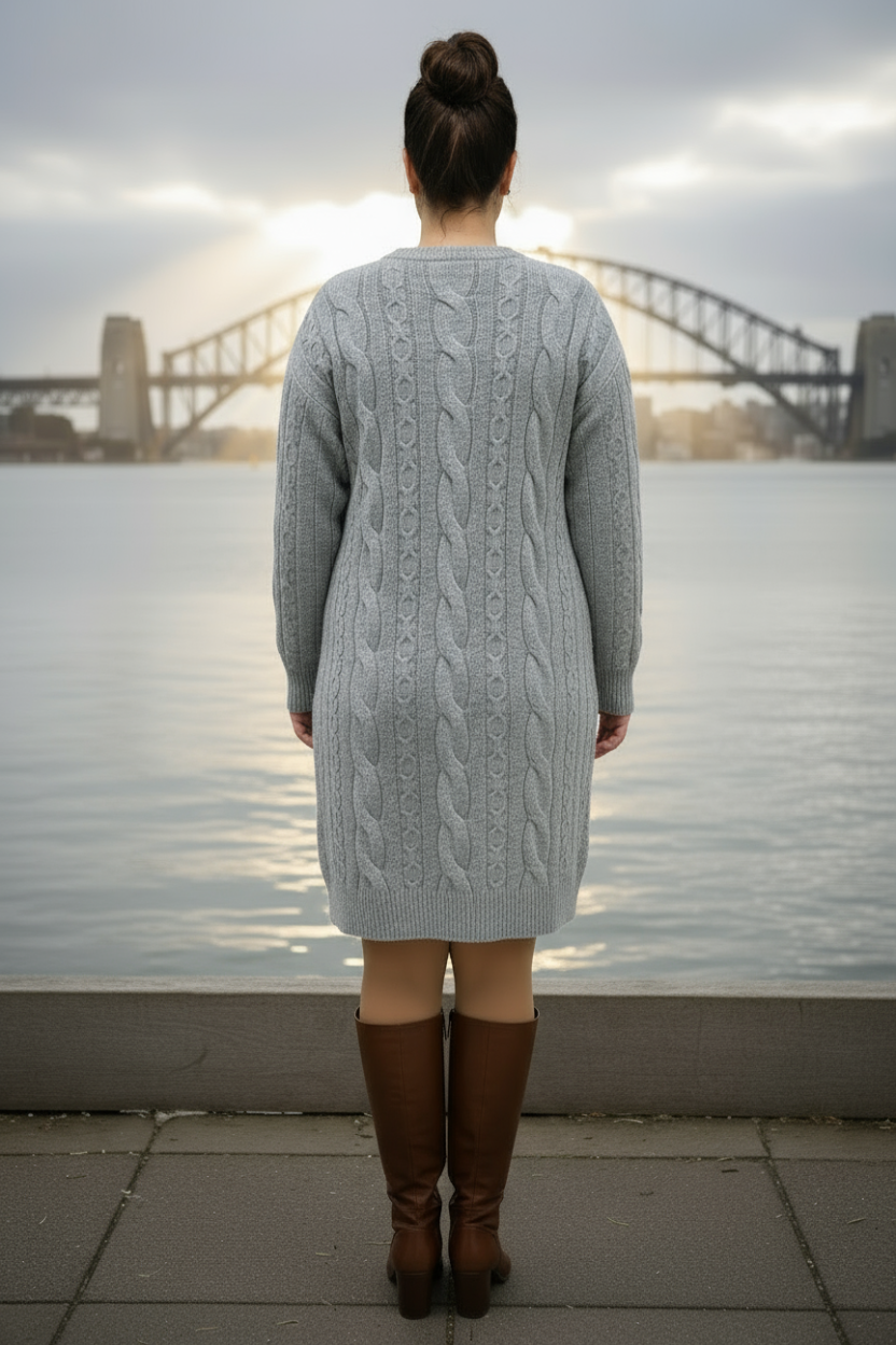 Person wearing a gray cable knit sweater and brown boots standing by a waterfront with a bridge in the background.