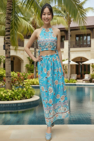 Woman in a blue floral dress standing by a pool with palm trees and a building in the background