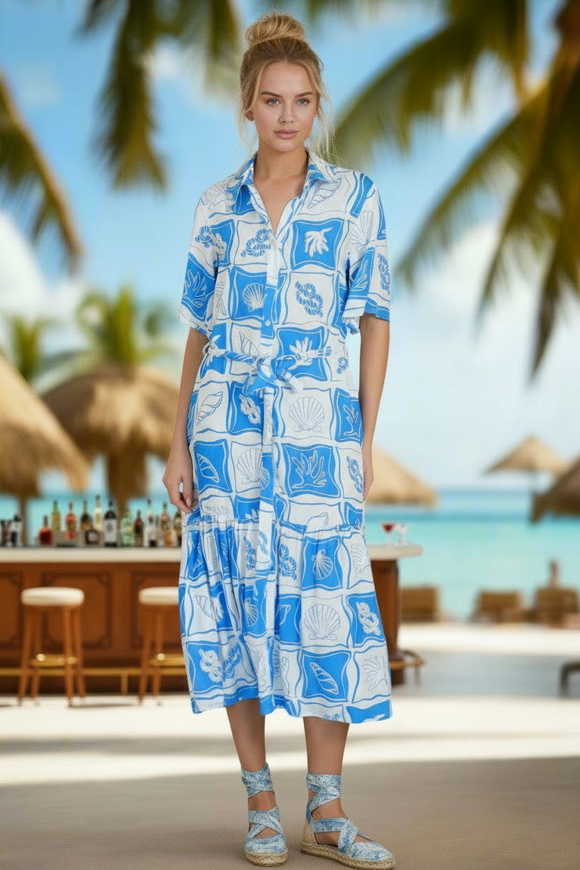 Woman in a blue and white patterned dress standing on a beach with palm trees and a thatched-roof hut in the background.