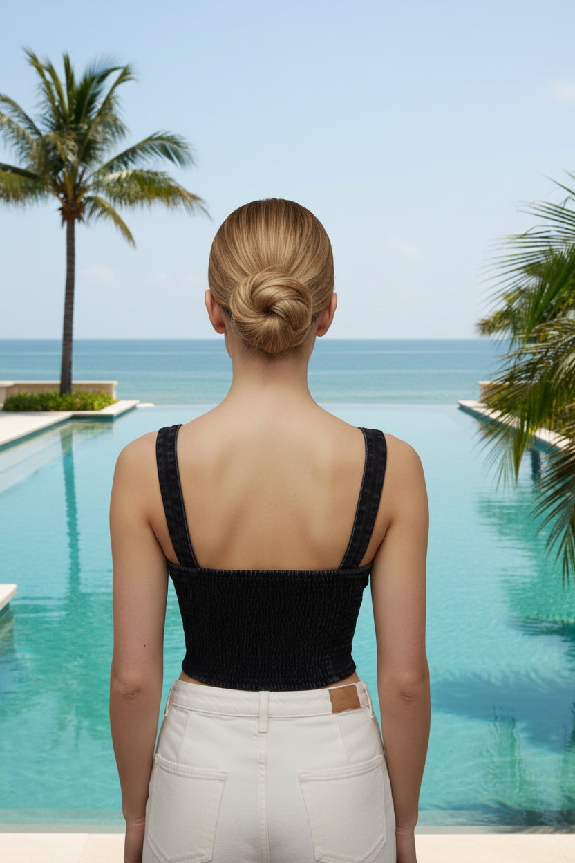 Woman wearing a black denim vest top with buttons on the front by a pool with palm trees in the background.