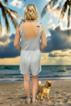 Woman in a light gray romper standing on a beach with a dog, palm trees, and ocean in the background.