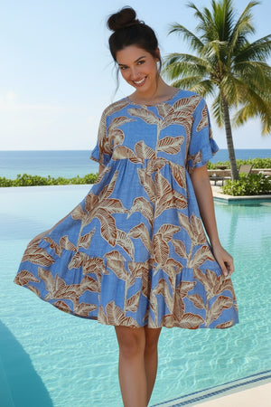 Woman wearing a blue dress with a leaf pattern standing against a white wall and stone wall.