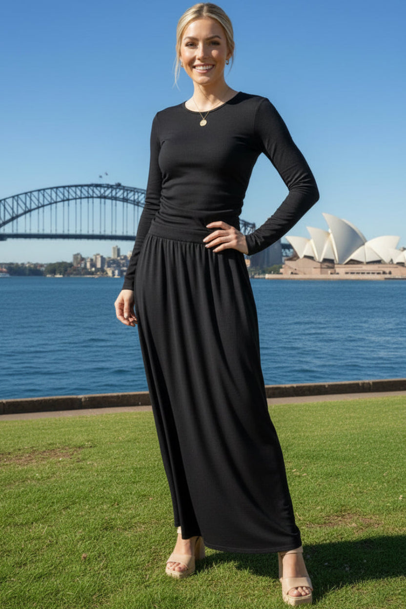 Woman in a long sleeve black maxi dress standing in front of the Sydney Harbour Bridge and Opera House.