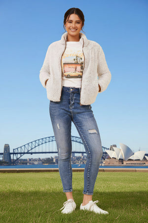 Woman wearing a white jacket, graphic t-shirt, and blue jeans standing in front of the Sydney Opera House and Harbour Bridge.