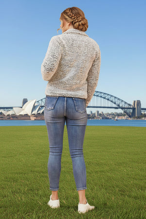 Person wearing a textured sweater and jeans standing on grass with a bridge and skyline in the background