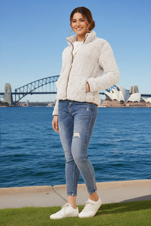 Woman wearing a light-colored jacket and jeans standing by a waterfront with a bridge in the background