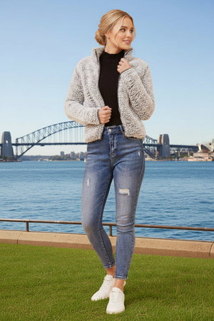 Woman wearing a gray jacket, black top, and blue jeans standing in front of a bridge over water.