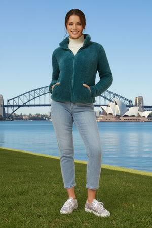 Woman in teal jacket and jeans standing in front of the Sydney Harbour Bridge and Opera House.