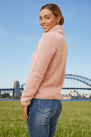 Woman wearing a pink sweater and blue jeans standing in a grassy field with a bridge in the background