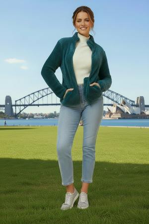 Woman in teal jacket and light jeans standing on grass with a bridge and water in the background
