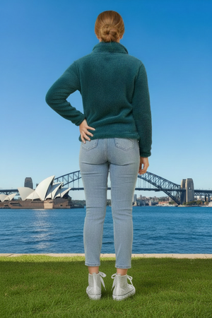 Person wearing a green jacket and light blue jeans standing on grass with the Sydney Opera House and Harbour Bridge in the background.