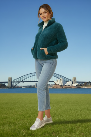 Woman in teal jacket and light blue jeans standing on grass with a bridge and Opera House in the background