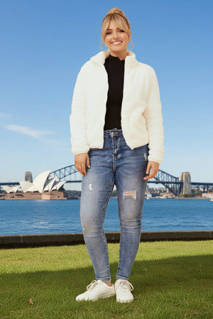 Woman in white jacket and jeans standing in front of the Sydney Opera House and Harbour Bridge.