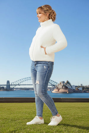 Woman in a white jacket and blue jeans standing on grass with a cityscape in the background