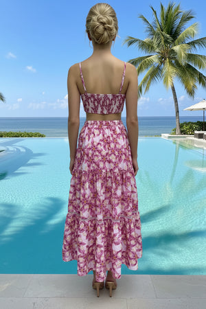 Woman in a pink floral maxi skirt and crop top standing by a pool with palm trees and ocean view.