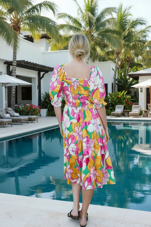 Woman wearing a colorful floral dress standing on a stone path with a white railing and plants in the background.