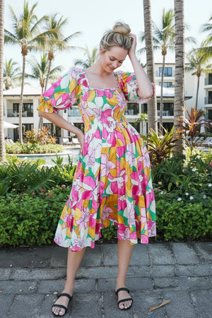 Woman in a colorful floral dress standing in a tropical setting with palm trees and a pool.