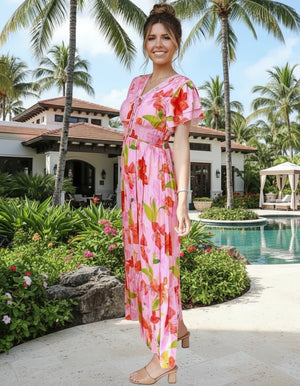 Woman in a pink floral dress standing in a tropical setting with palm trees and a pool.