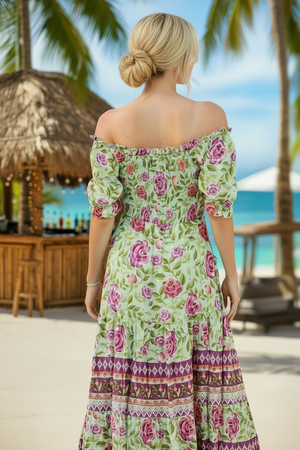 Woman in a floral dress standing on a tropical beach with palm trees and a bar in the background.